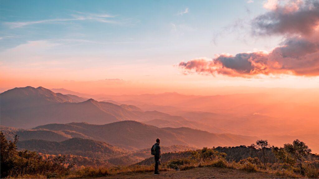 Person standing in the distance on a mountaintop with a background of other mountains and a red and blue sunset.