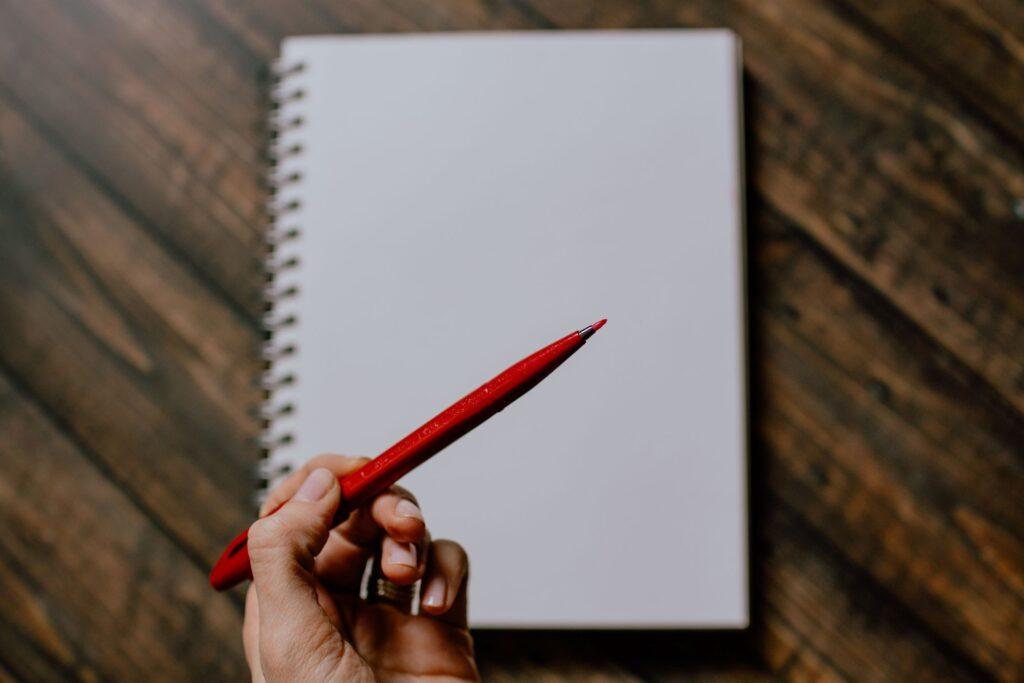 Person's hand holds a red pen above a blurred white notebook, which sits on a wooden desk.