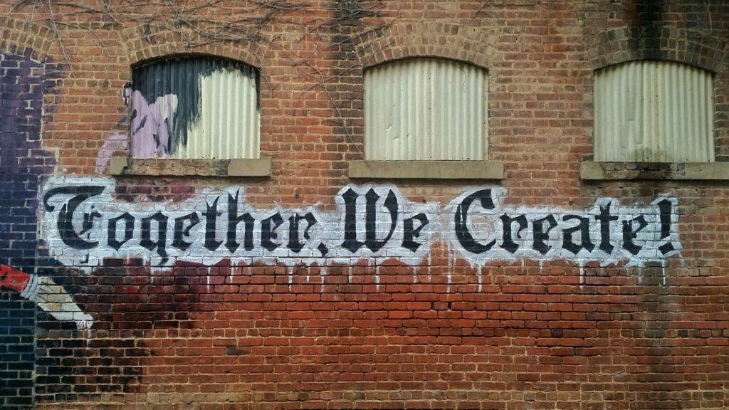 Brick wall featuring graffiti in old-fashioned lettering that reads, "Together We Wreate!"