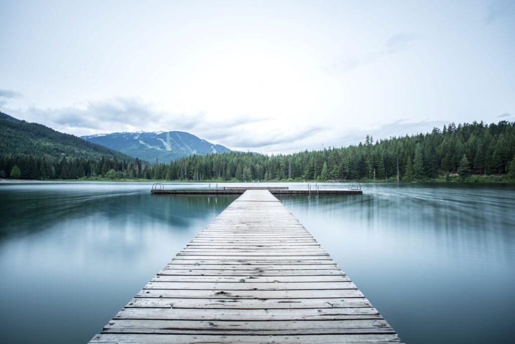 Open lake with a wooden dock leading into the center and a background of forests and mountains.
