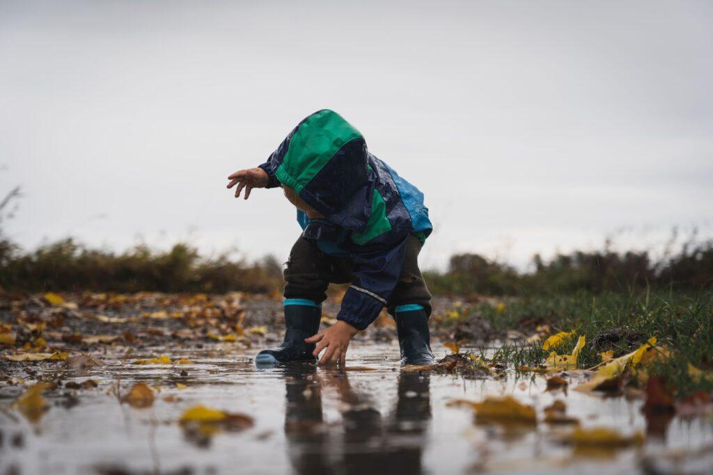 Child in a blue and green raincoat and rubber boots stands in a puddle and reaches toward the water.